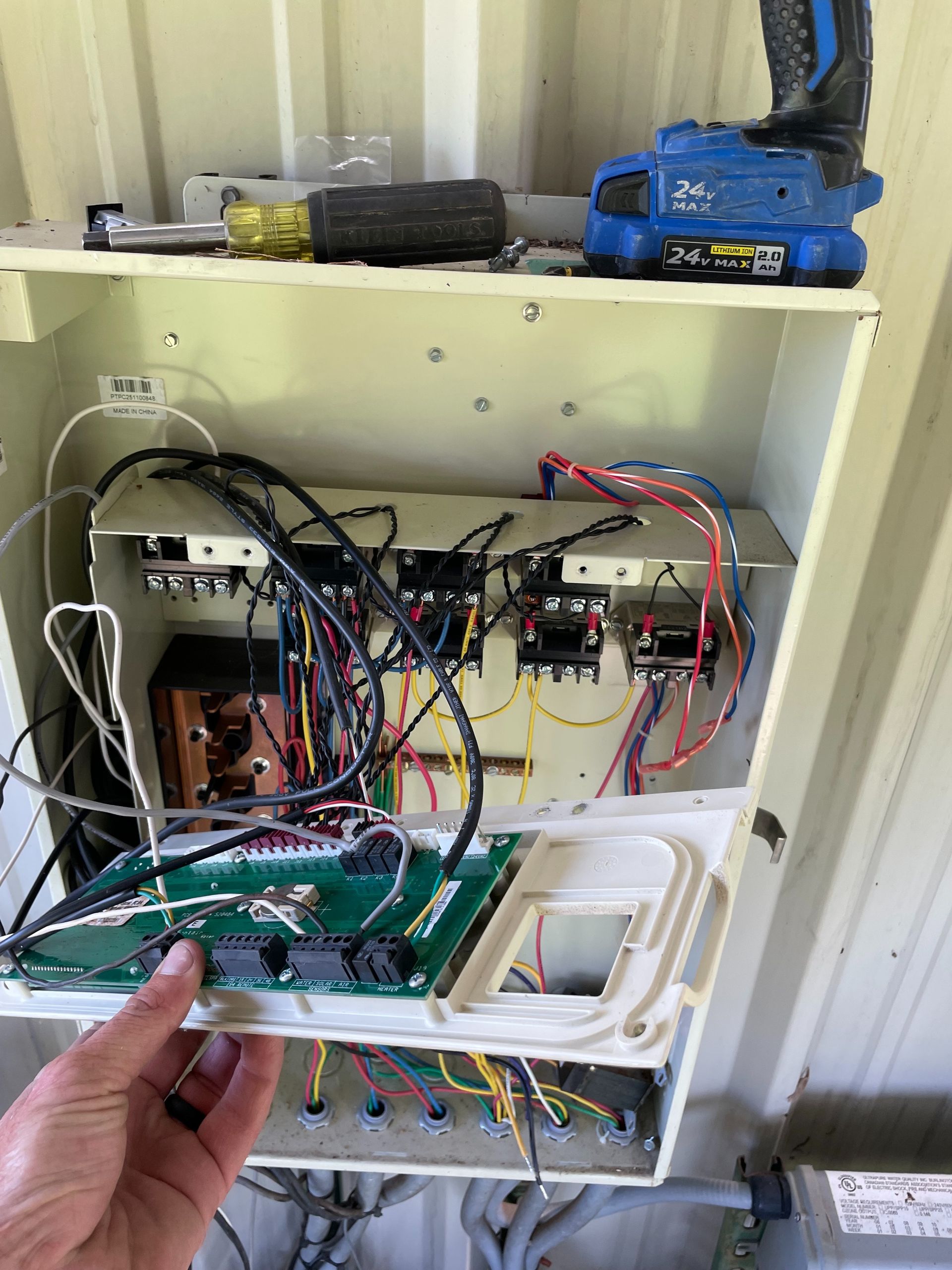 Hand holding a green circuit board in front of an open electronics cabinet with tangled wires and blue equipment