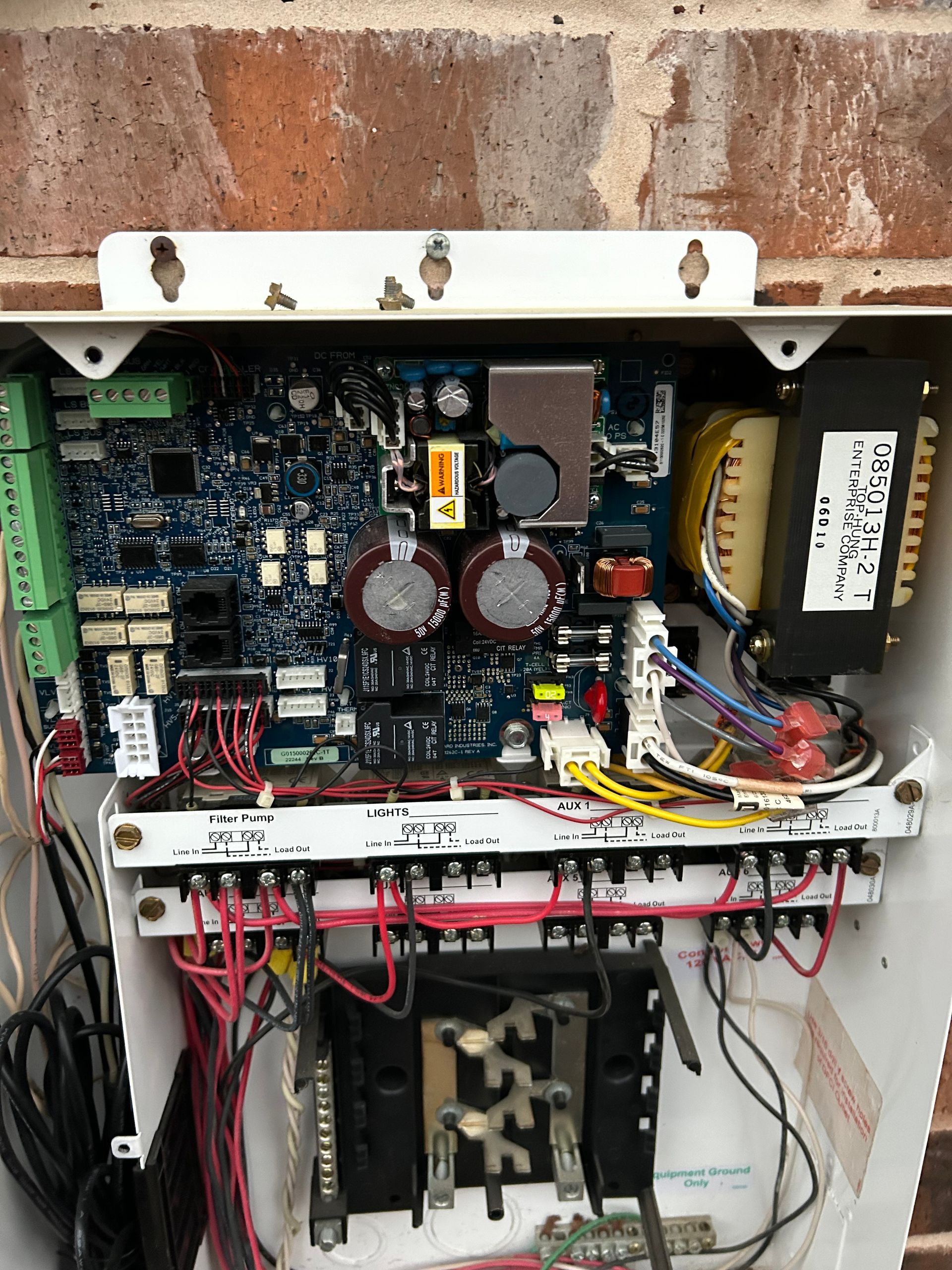 Open electrical control panel with circuit boards, wires, and components mounted on a brick wall.