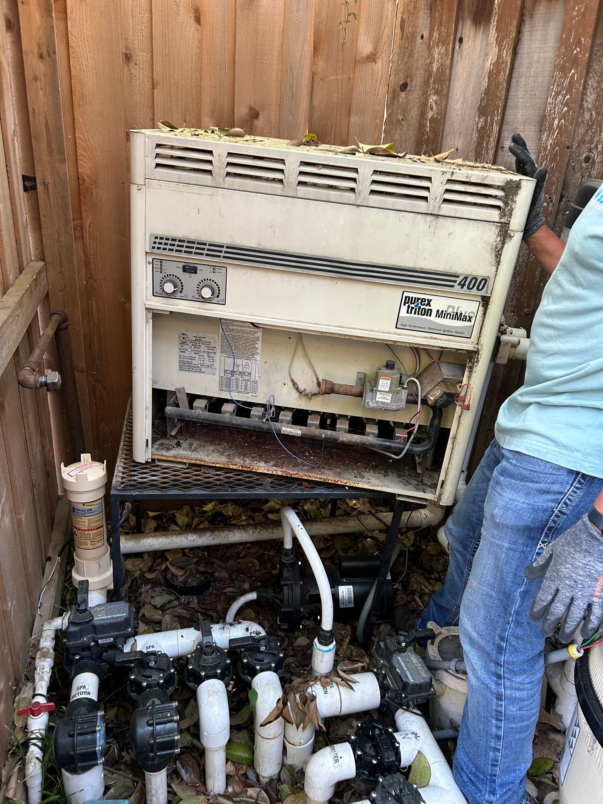 Open electrical panel mounted on a wooden fence with exposed wires and bottles on the ground beneath it
