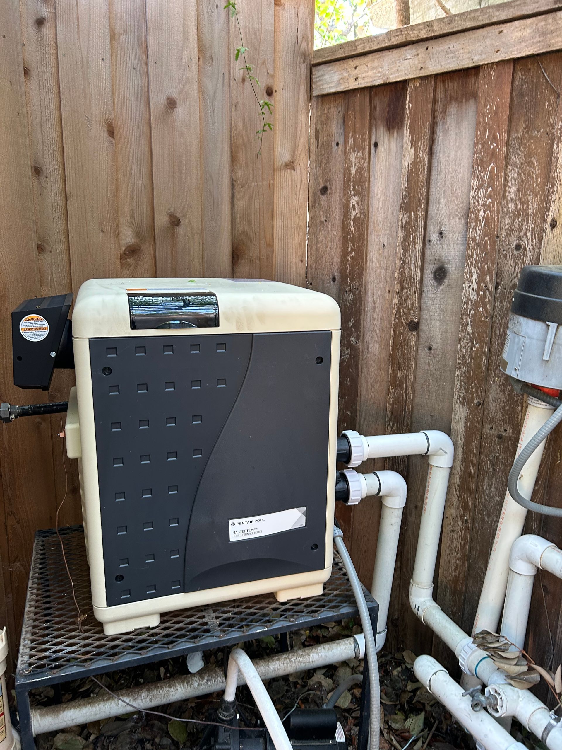 Outdoor HVAC unit beside a wooden fence, with white PVC pipes and a gravel ground.