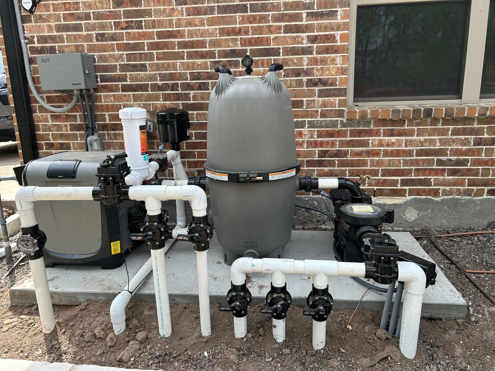 Outdoor pool filtration and pump equipment with white PVC pipes beside a brick wall.