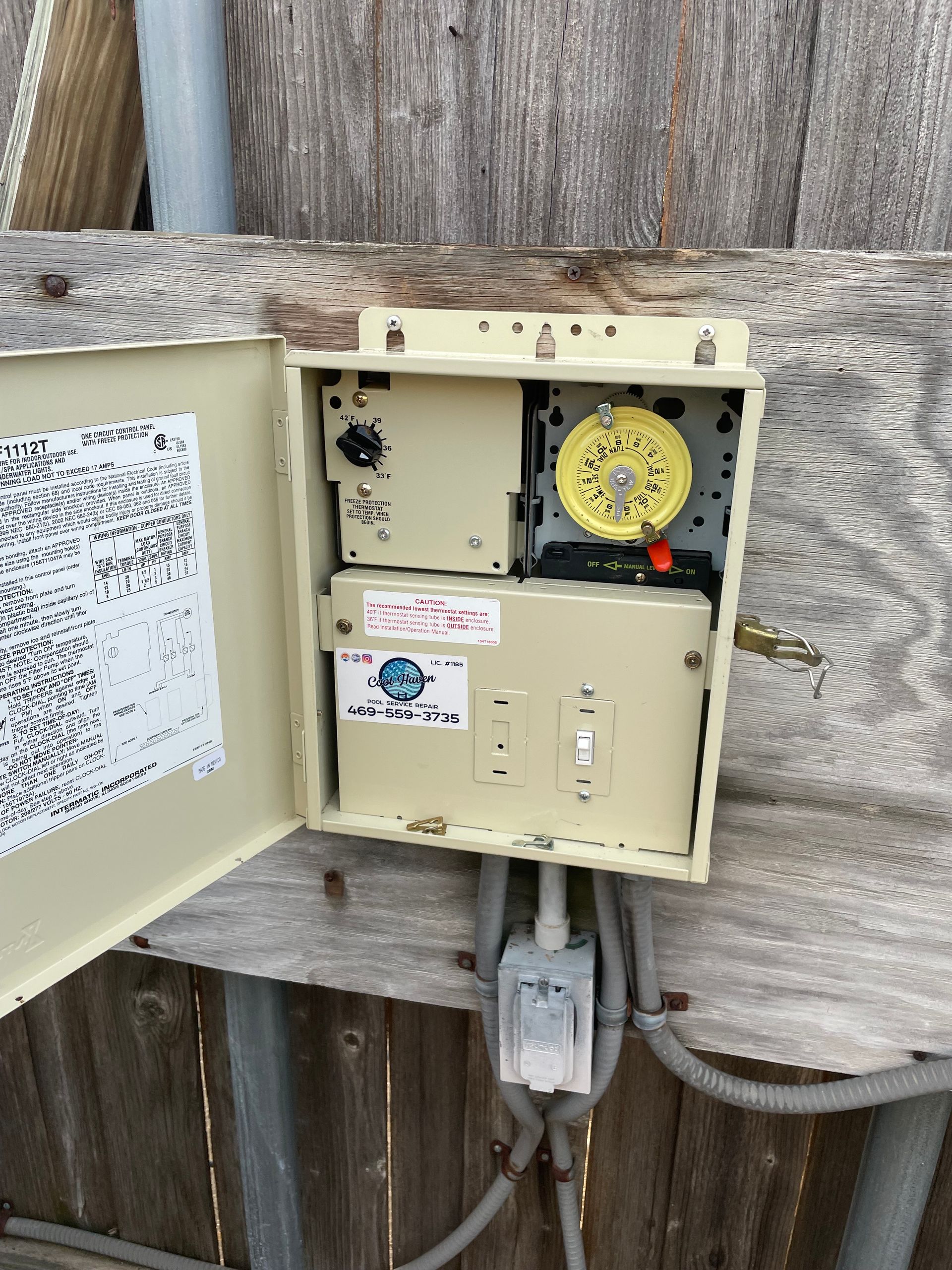 Open electrical control box with yellow timer mounted on a wooden fence outdoors.