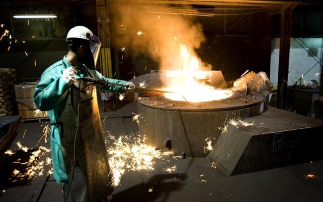 Worker in green coat tending a blazing metal furnace in an industrial workshop