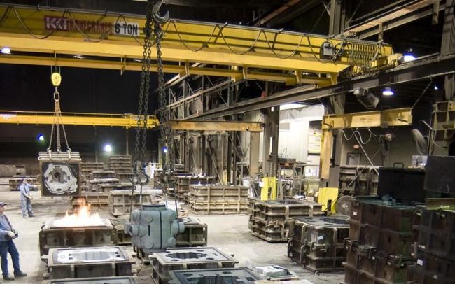 Industrial factory floor with large yellow overhead crane, machinery, stacked metal parts, and workers near a welding flame