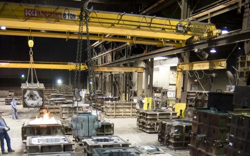 Industrial factory floor with yellow overhead cranes, machinery, and workers handling metal parts