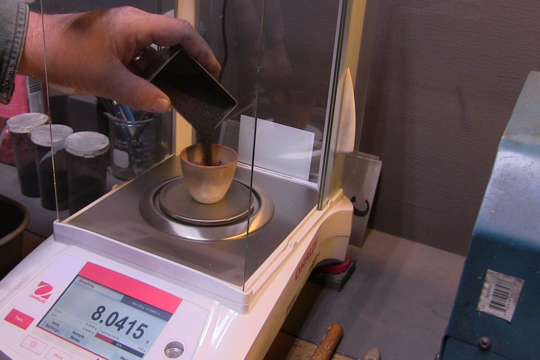 Hand pouring something into a small cup on a digital scale in a lab or workshop.