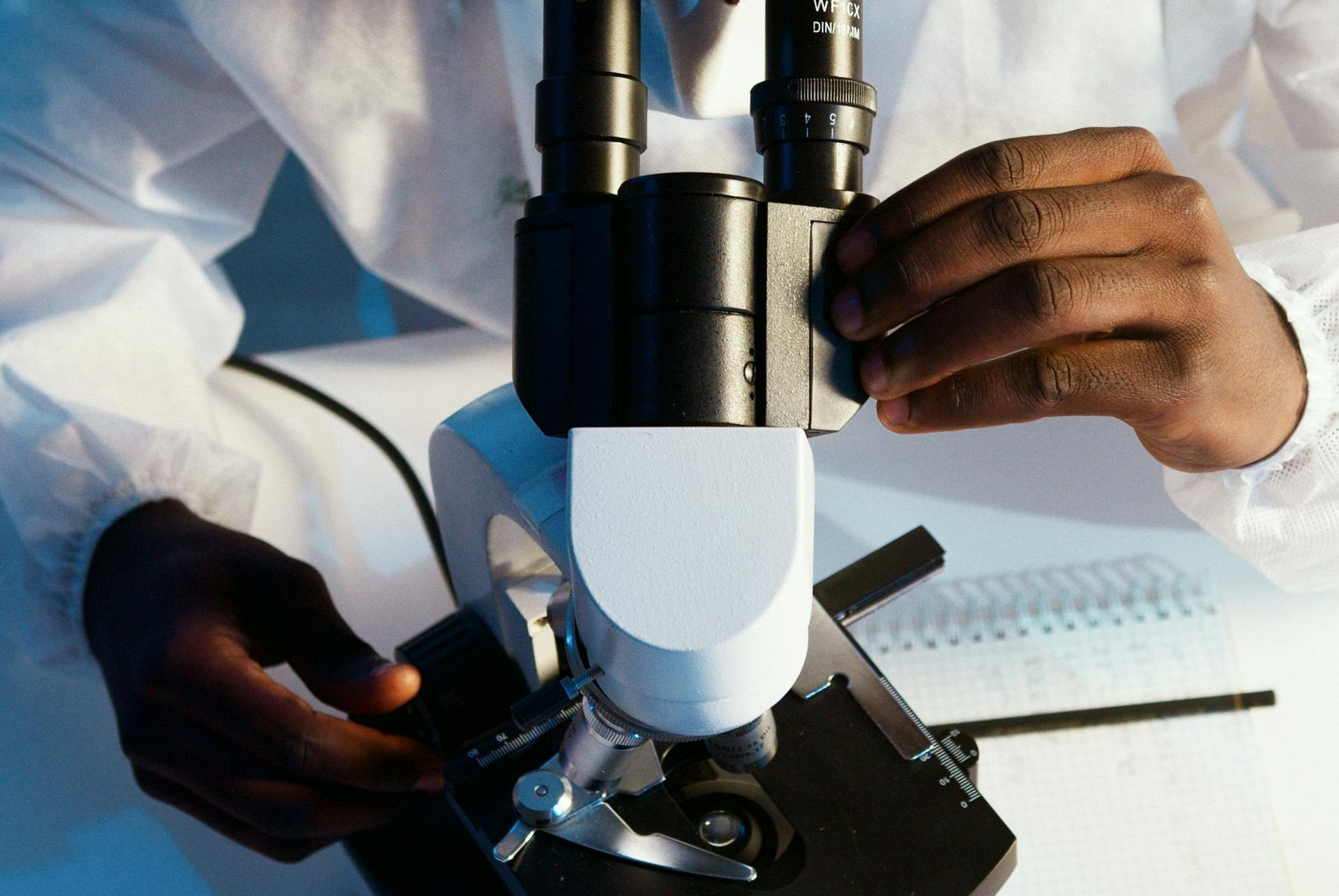 Hands adjusting a microscope in a lab setting, with white lab coat and notebook nearby