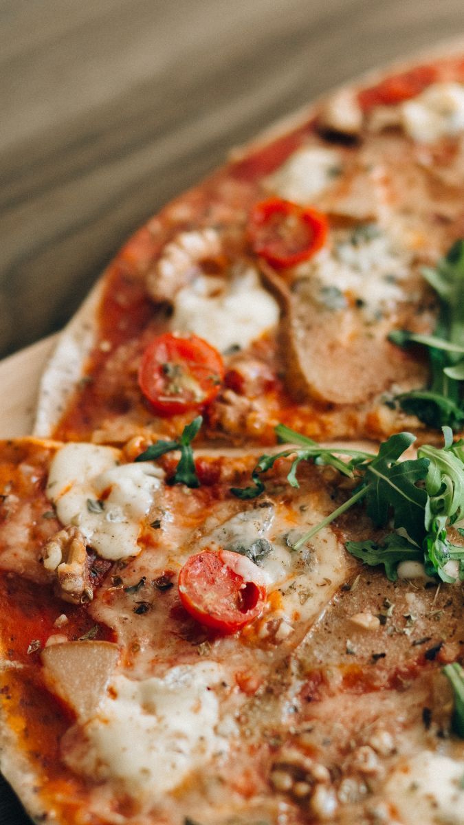 Nahaufnahme einer Pizza mit geschmolzenem Käse, Kirschtomaten, Kräutern und Rucola auf einem Holztisch