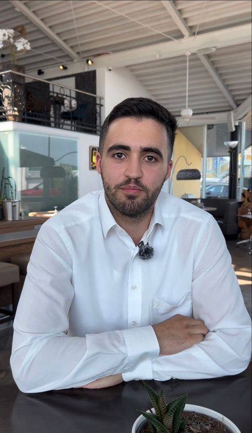 Ein lächelnder Mann mit Brille und blauer Jacke sitzt drinnen an einem Café-Tresen; hinter ihm ist ein „Bombay“-Schild zu sehen.