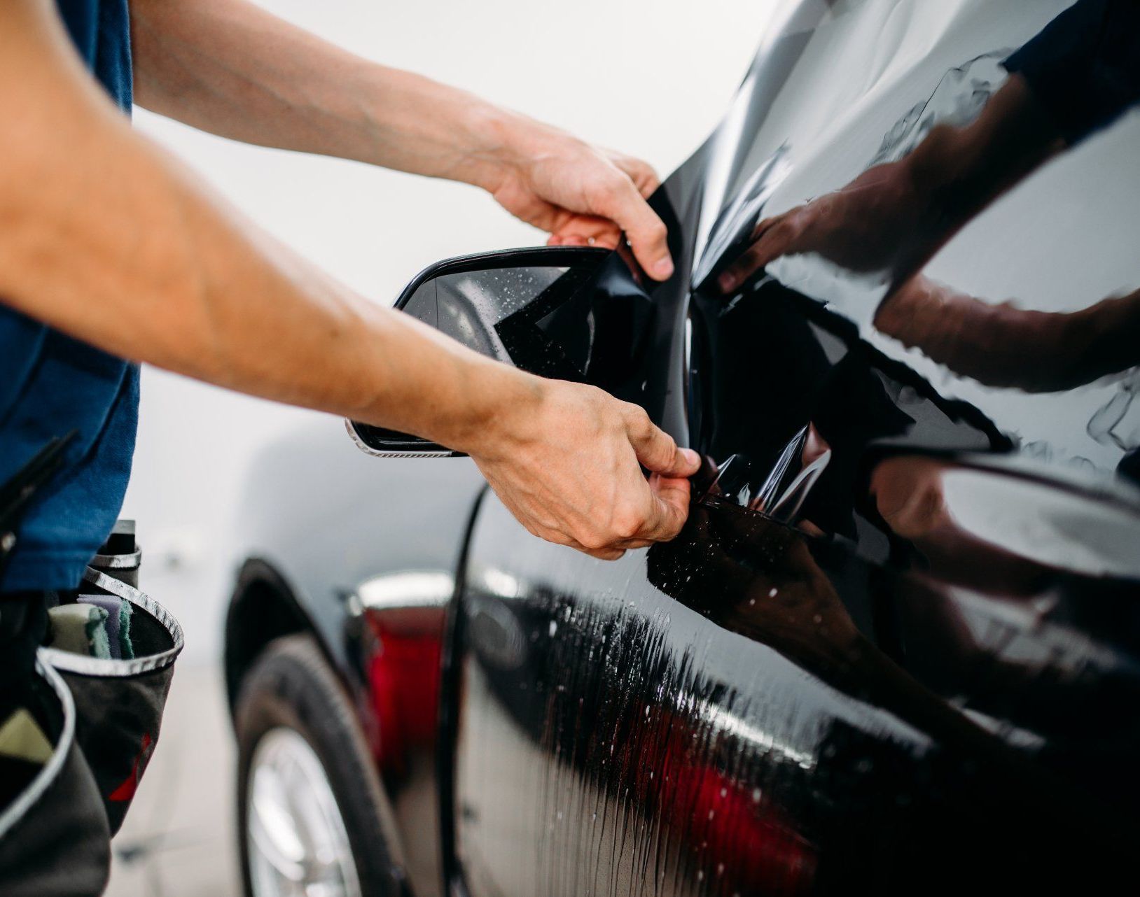 Hands installing a clear protective film on a black car door in a detailing shop