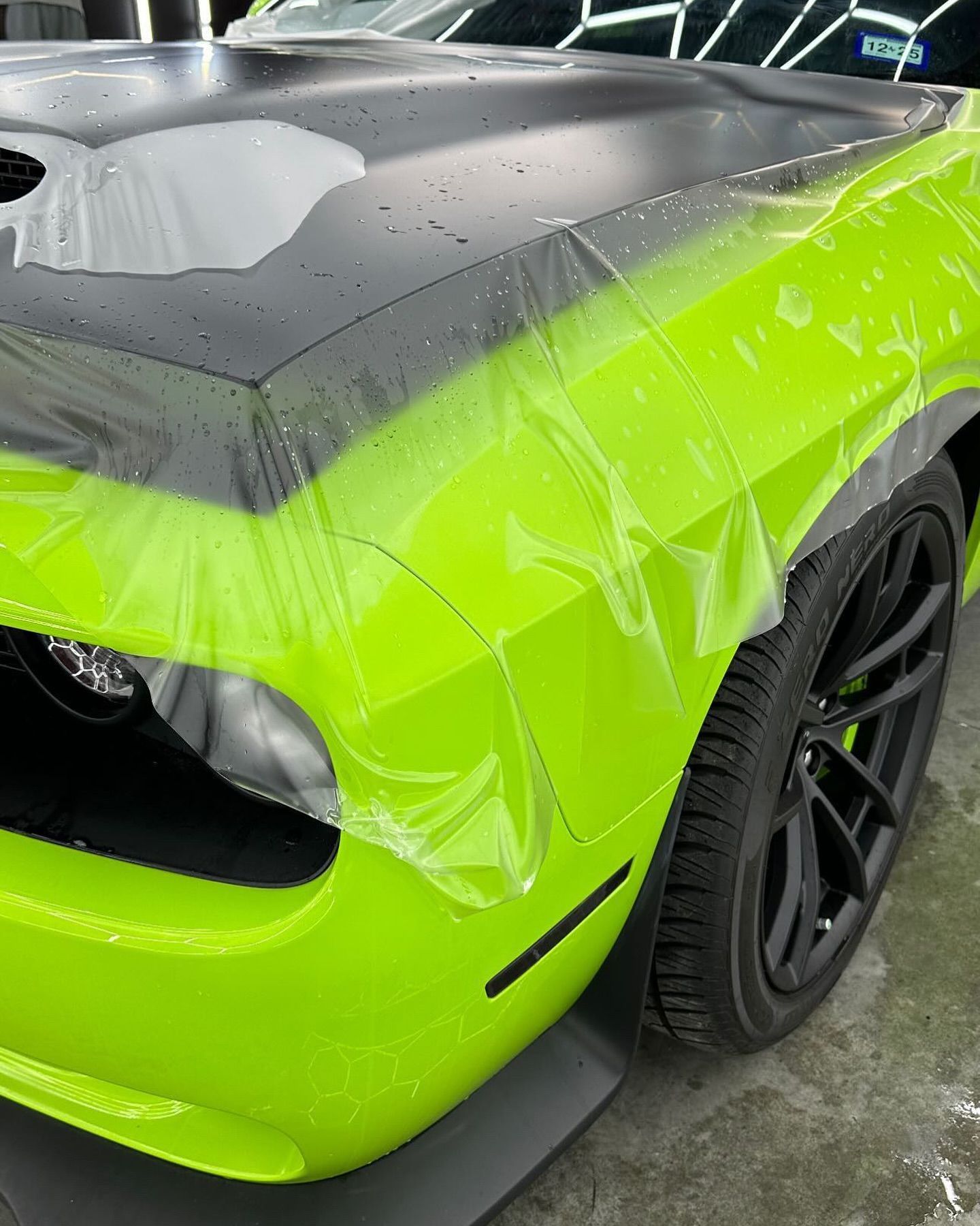 Neon green sports car wrapped in clear plastic with a black hood and black wheels in a garage