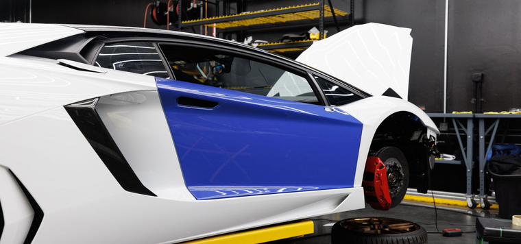 White sports car with a blue door panel in a workshop, front wheel removed on a lift, being repaired