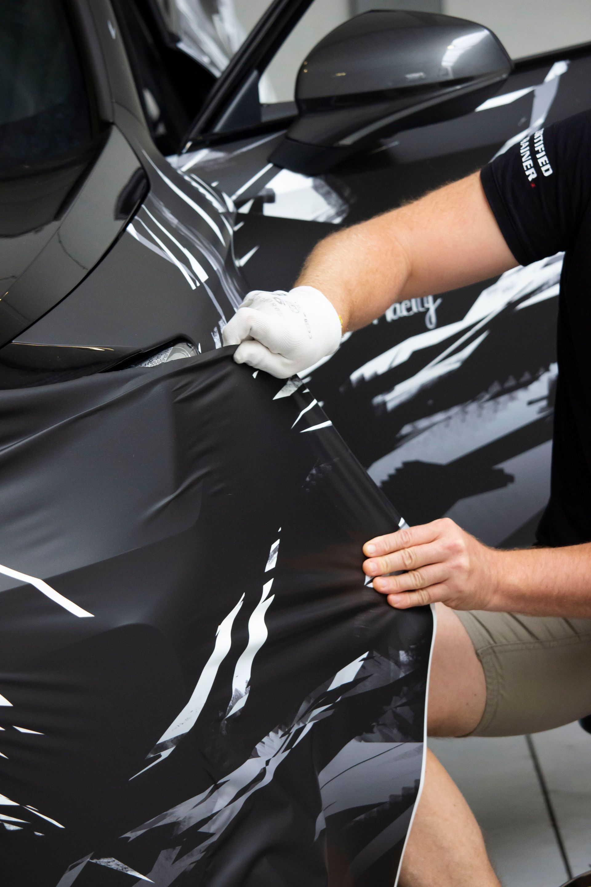 Person applying black vinyl wrap to a car hood with a squeegee and gloved hands