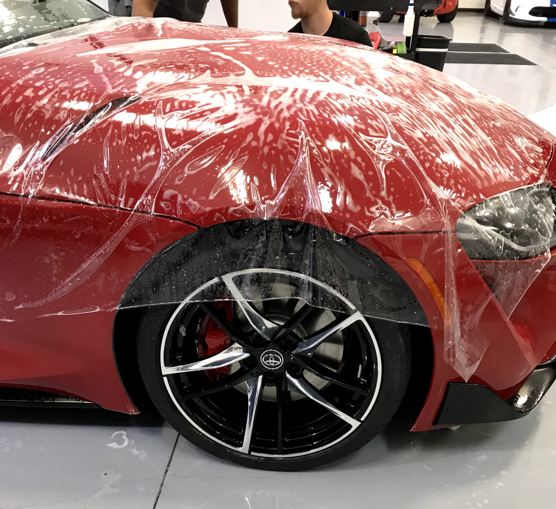 Red sports car front fender covered in clear protective plastic wrap at an auto show