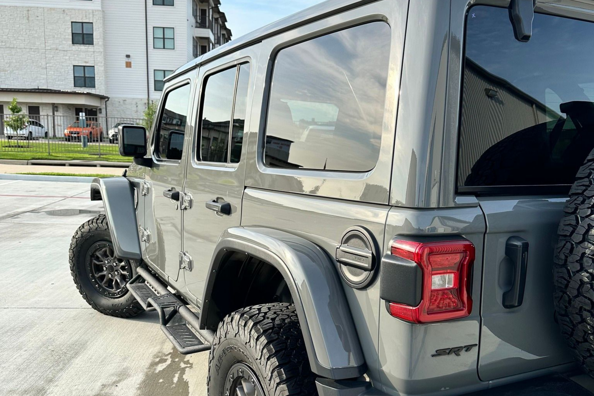 Gray Jeep Wrangler parked outside a building, rear side view with black off-road tires.