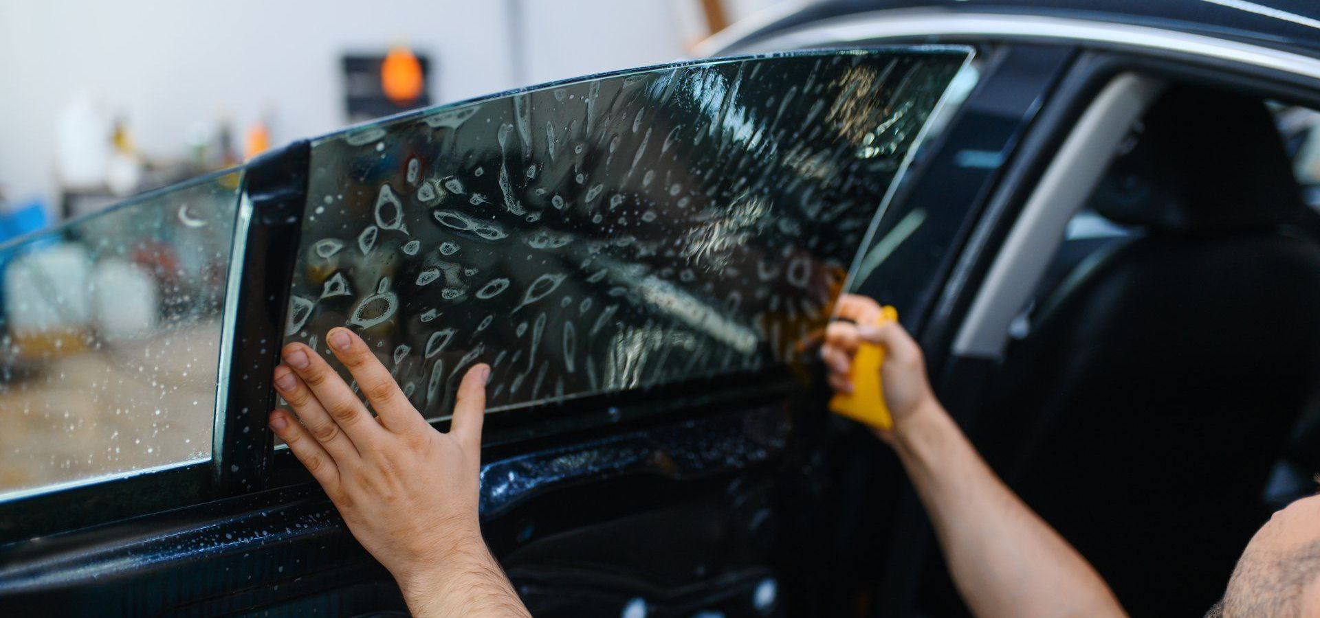 Hands tinting a car window with dark film outside an auto shop