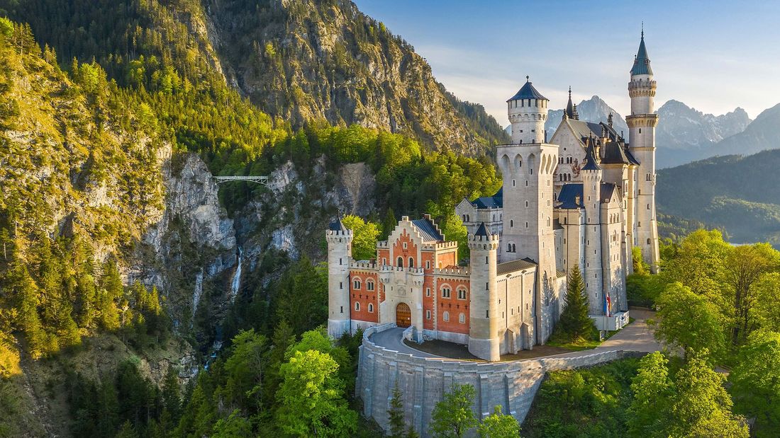 Fairytale castle with white towers on a forested cliff, surrounded by mountains at sunset