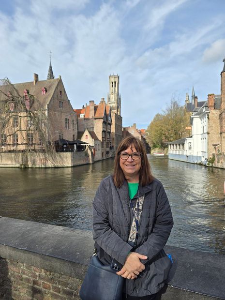 Woman standing by a canal in a historic European town, with old buildings and a bridge in the background.