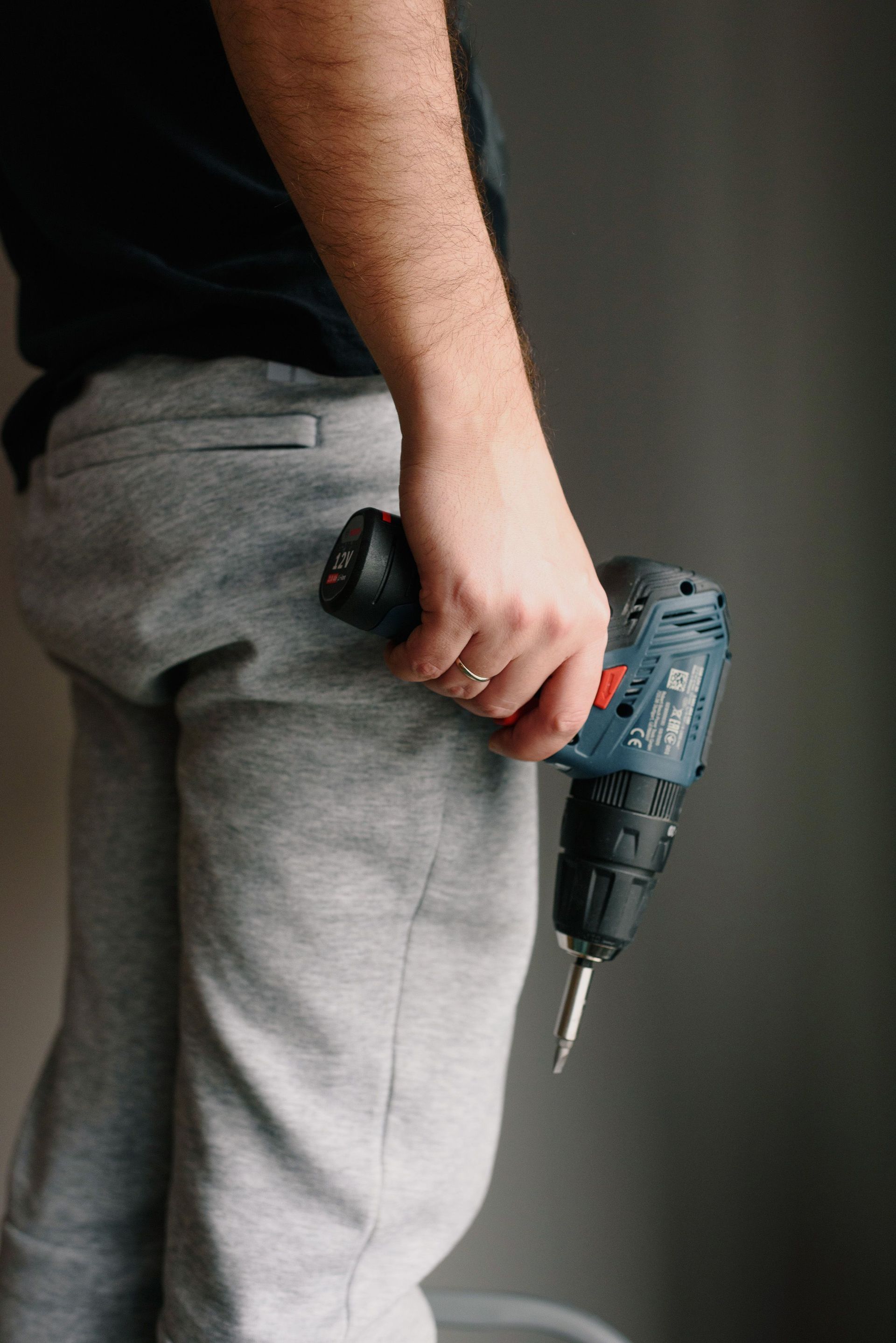 Person holding a gray power drill at their side, wearing gray pants indoors