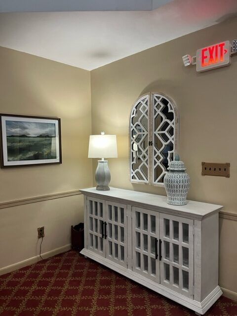 Hallway corner with white console table, lamp, mirror, framed art, and red carpet under an EXIT sign