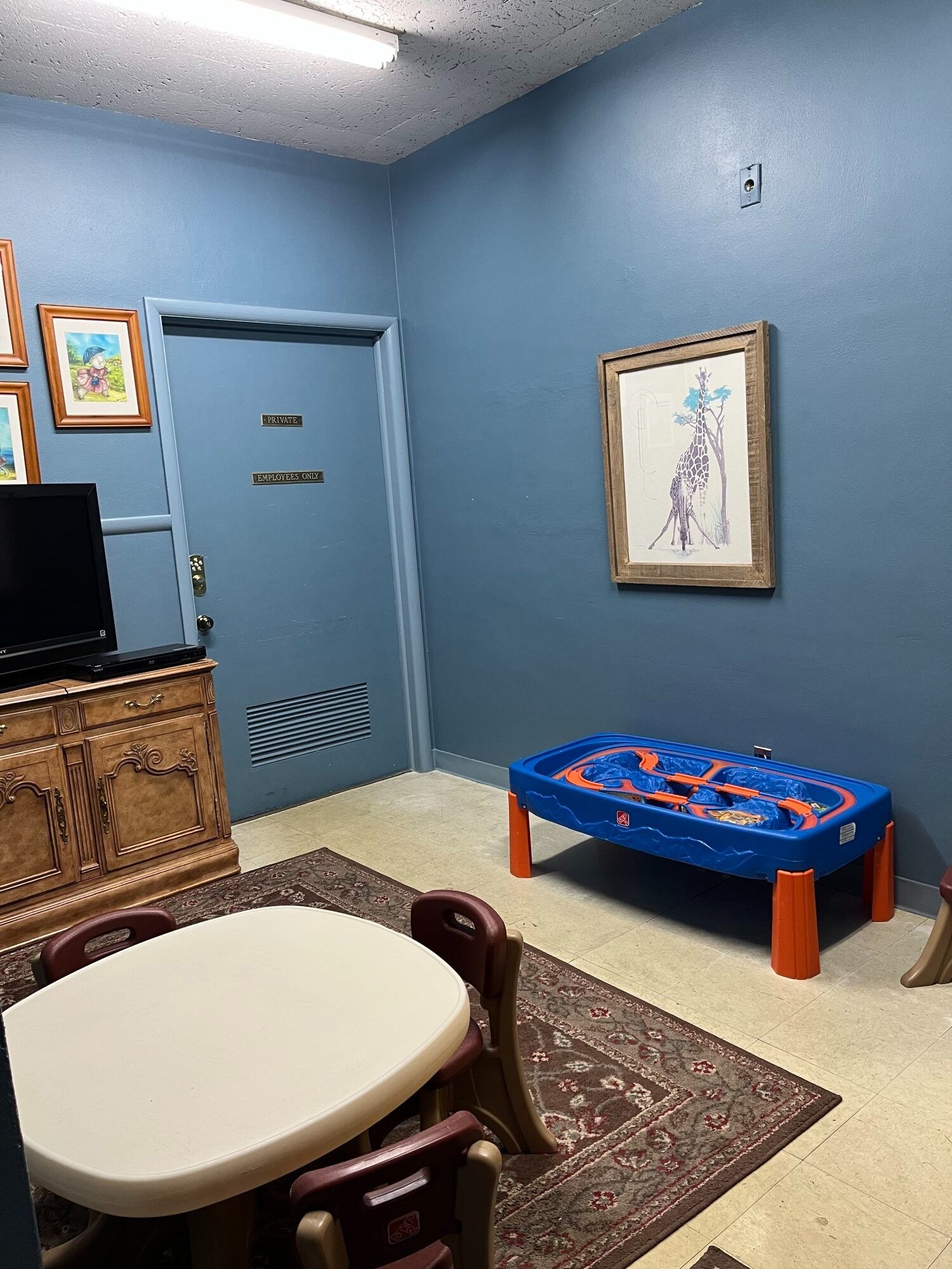 Blue waiting room with two small tables, a patterned rug, framed art, a TV, and a blue door.
