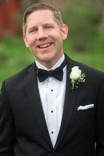 Smiling man in a black tuxedo with a white boutonniere outdoors, wearing a bow tie.