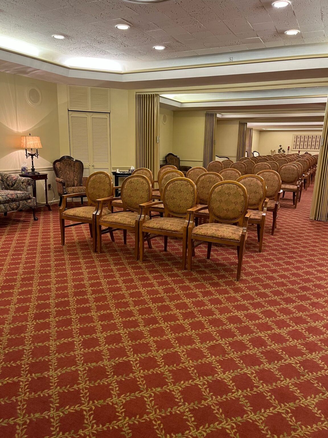 Empty banquet room with rows of upholstered chairs on a red patterned carpet.