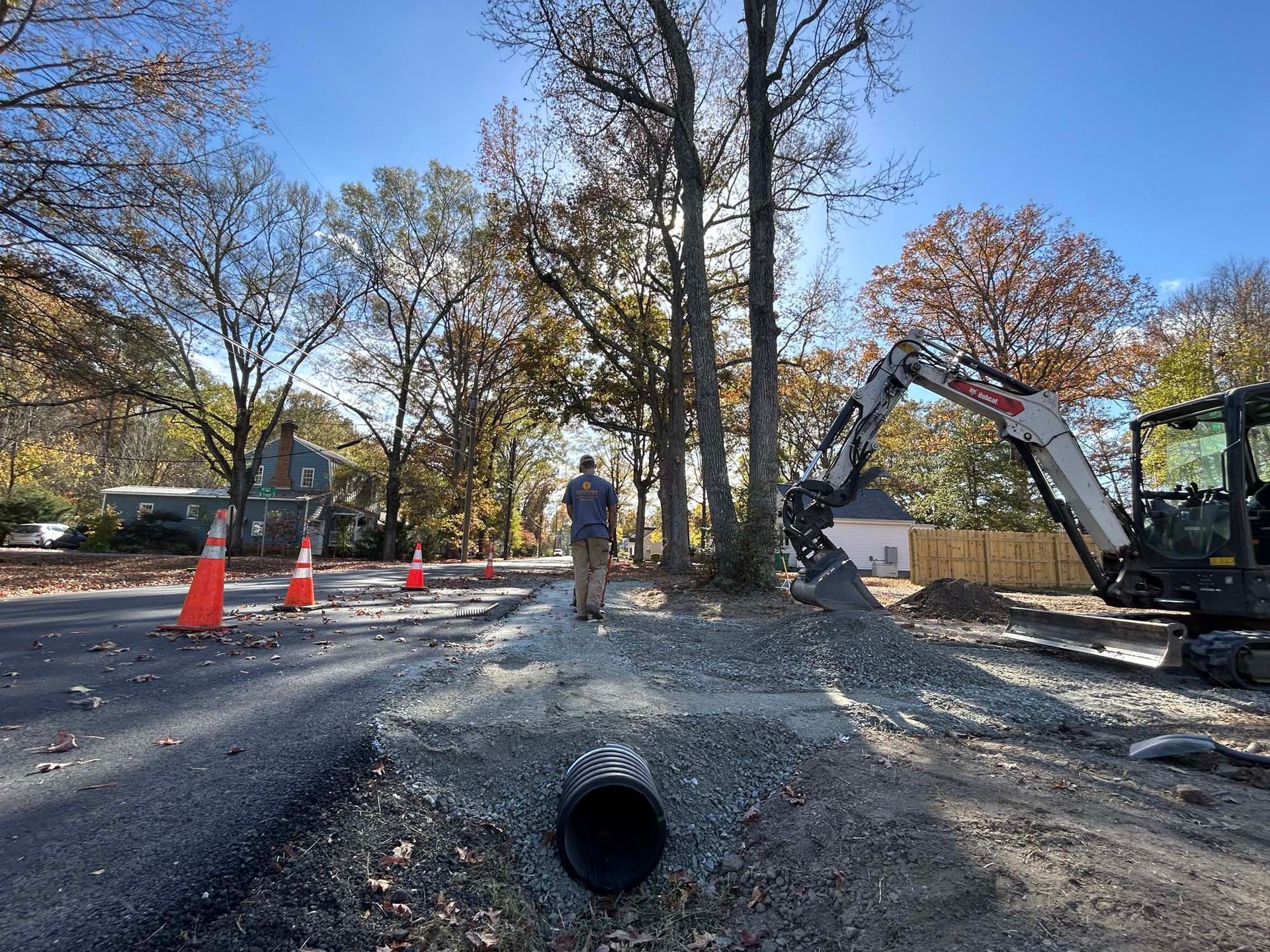 Construction site with an excavator, worker, traffic cones, and a large pipe in gravel roadwork area
