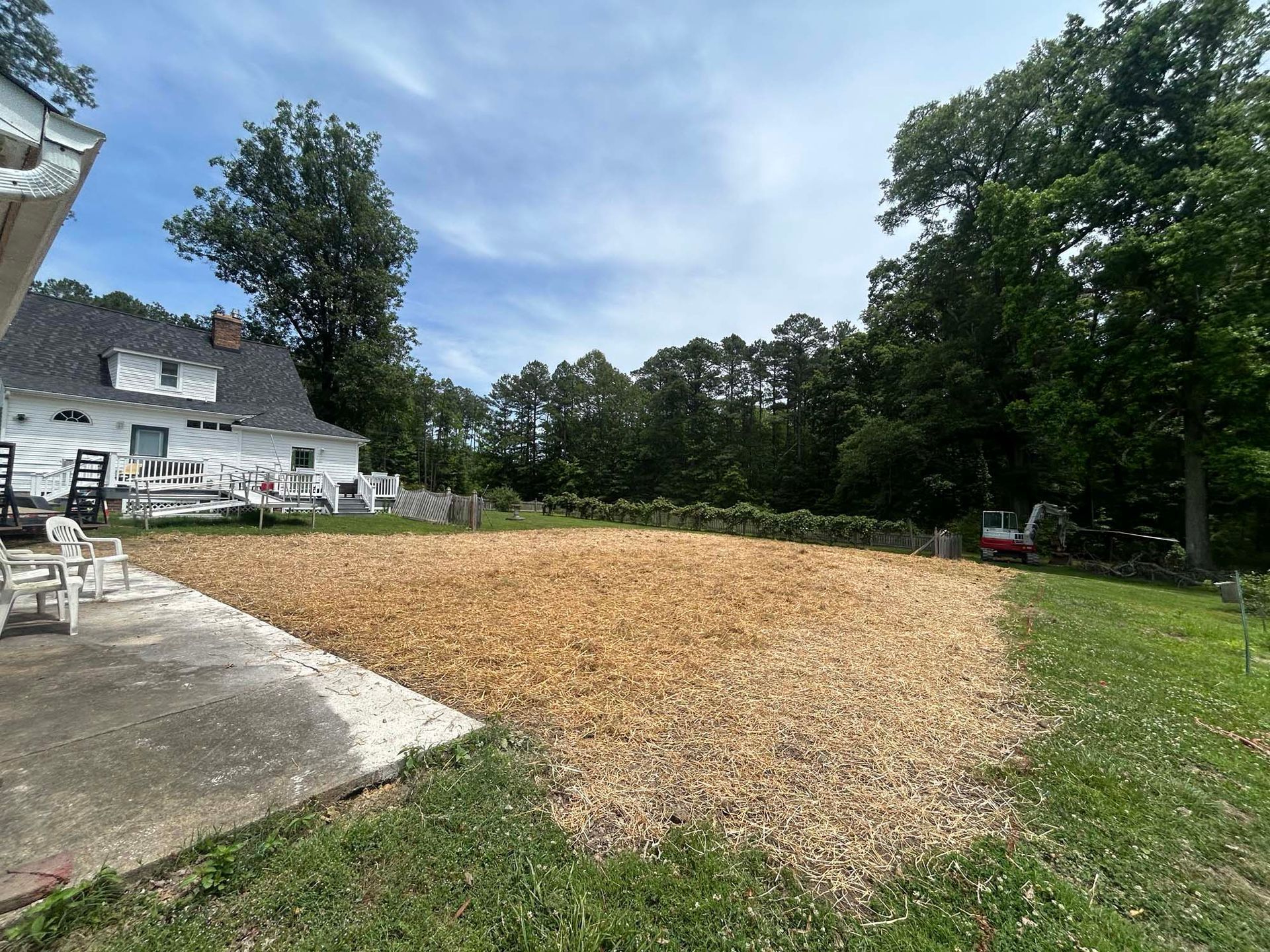 Backyard with a large wood-chip patch beside a patio, house, and trees under a blue sky