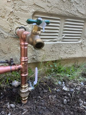 A brass outdoor faucet mounted on copper piping against an exterior stucco wall with a vent.