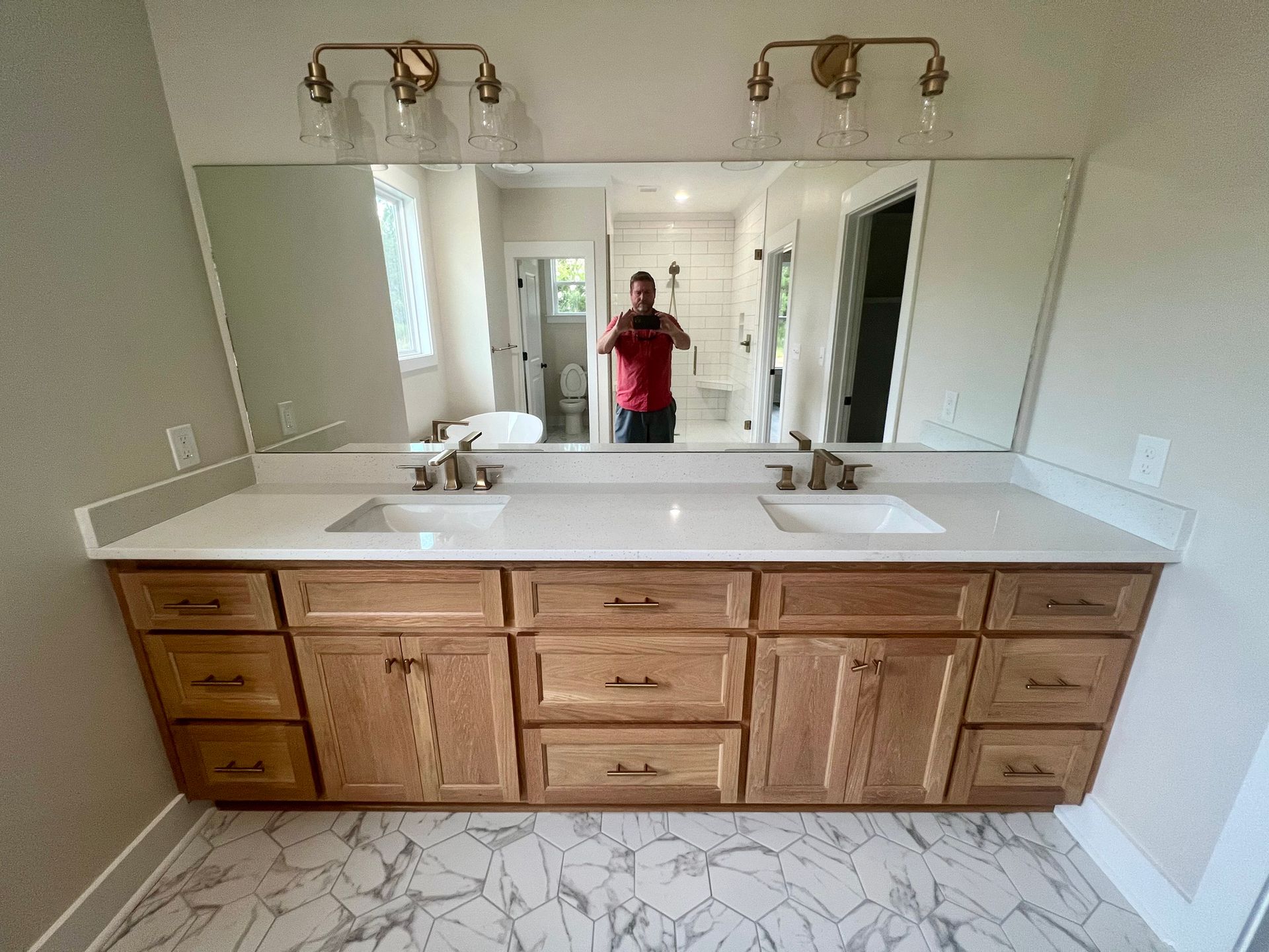 Bathroom vanity with oak cabinets, white countertop, two sinks, mirrors, and lights; person taking a picture.