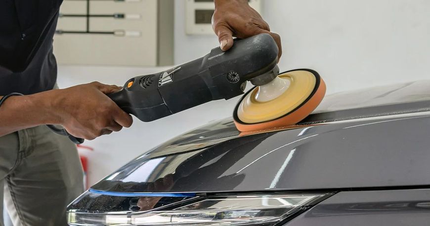 Persona che lucida il cofano scuro di un'auto con una lucidatrice elettrica e un tampone arancione.