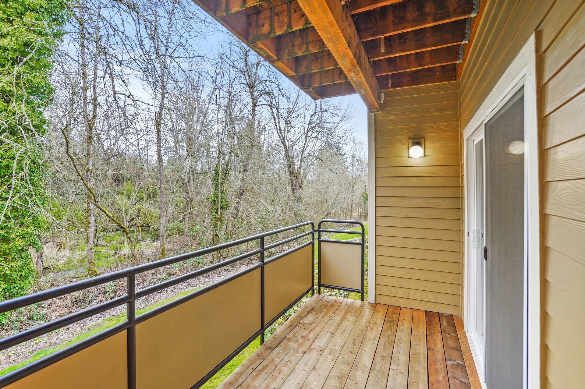 Covered balcony with wood decking, tan siding, black metal railing, sliding glass door, and wooded views beyond the railing.