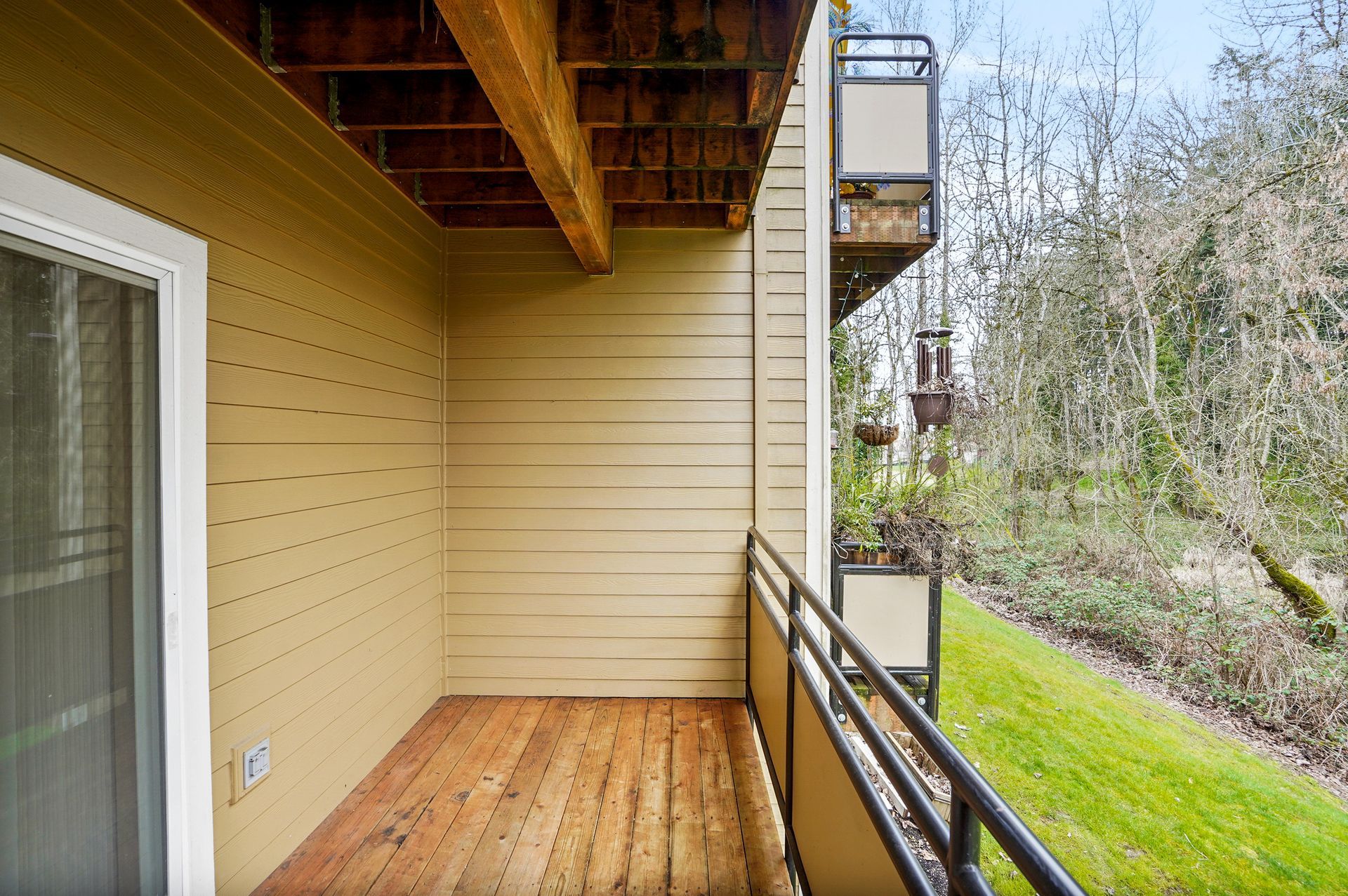 Covered balcony with wood decking, beige siding, black metal railing, and views of greenery and neighboring balconies.