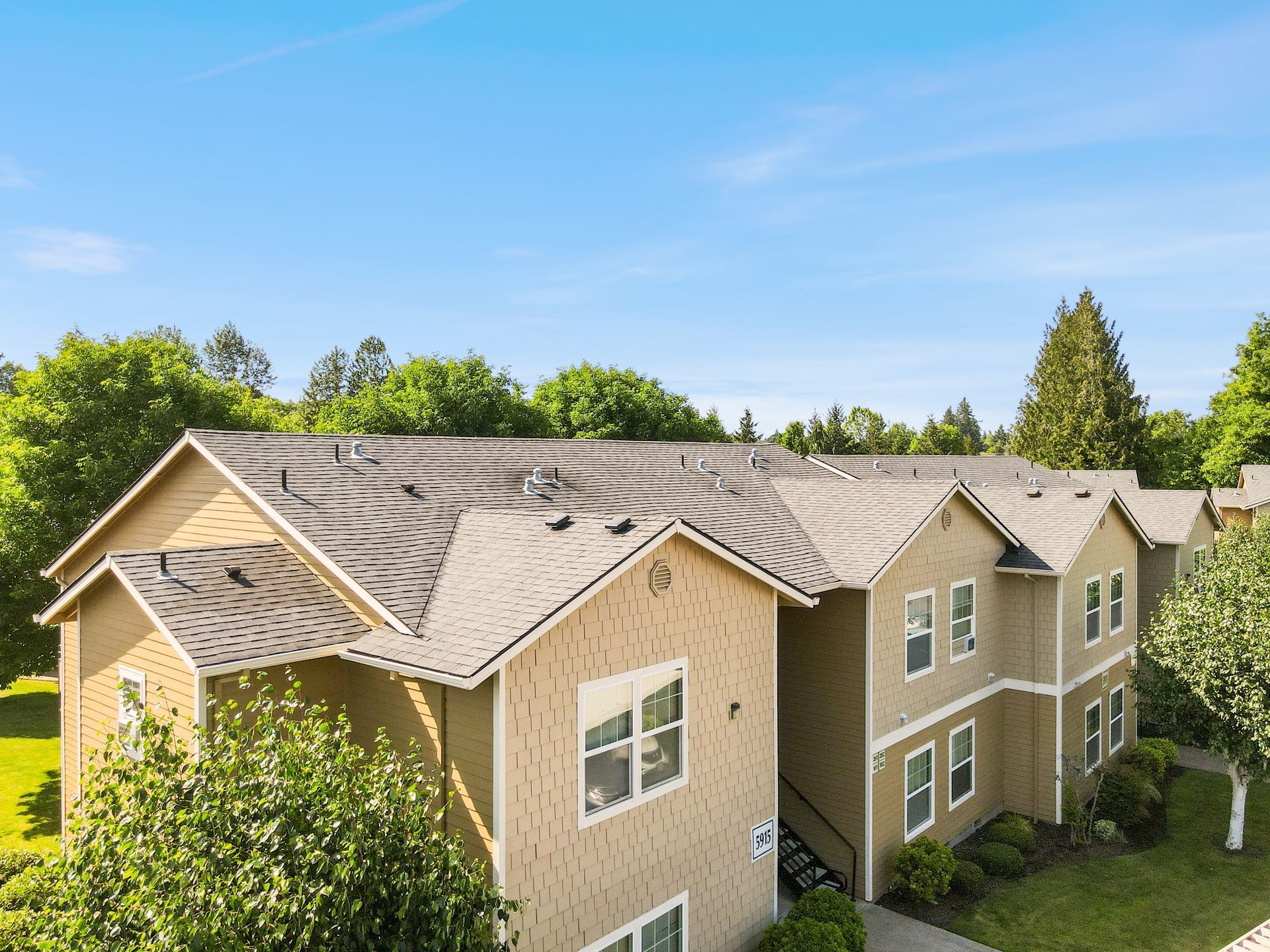 Exterior view of tan apartment buildings with pitched roofs, surrounded by trees and greenery under a clear blue sky.