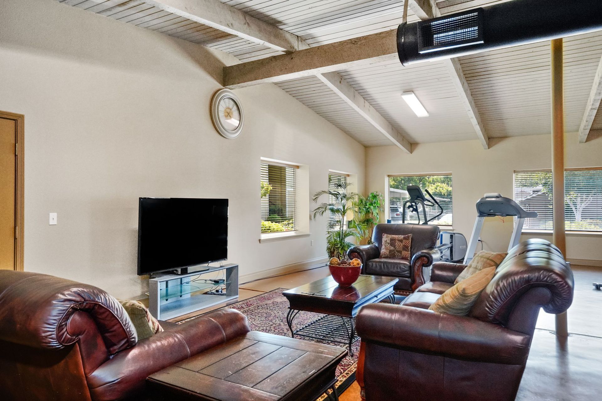 Community lounge with leather seating, TV, coffee table, exposed beams, and fitness equipment visible in the background.