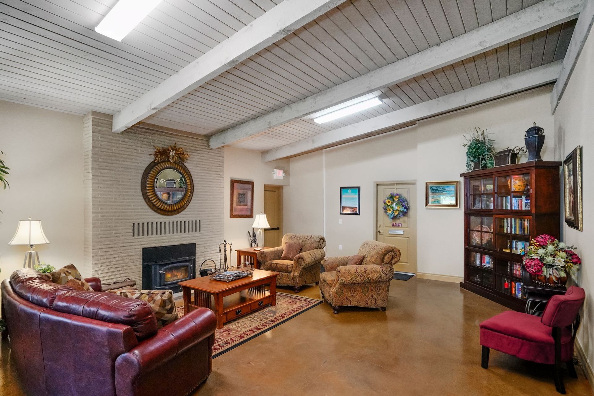 Community lounge with fireplace, leather and upholstered seating, coffee table, bookshelves, and exposed ceiling beams.