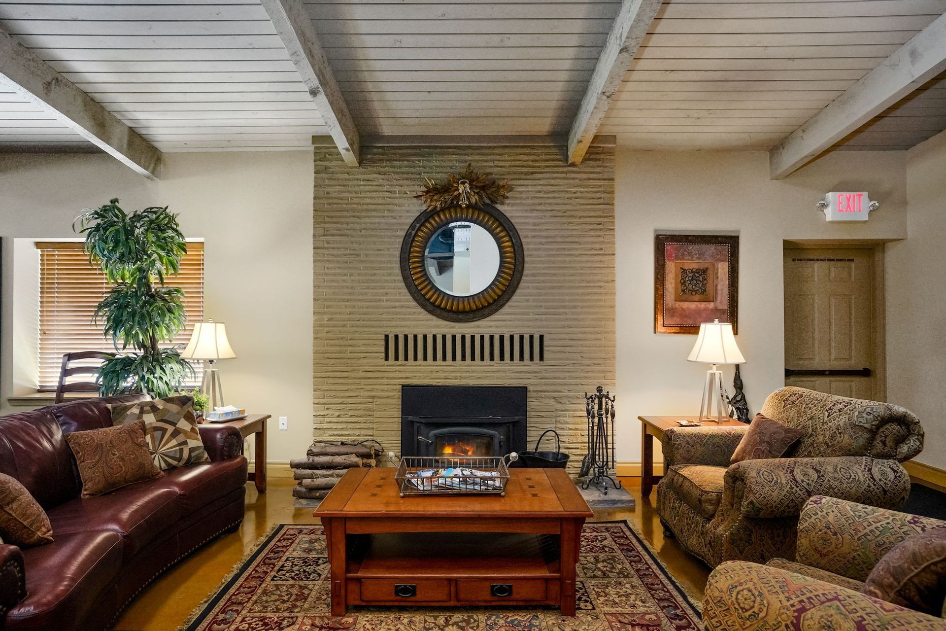 Community lounge with fireplace, sofas and armchairs, coffee table on an area rug, wall mirror, and exposed ceiling beams.