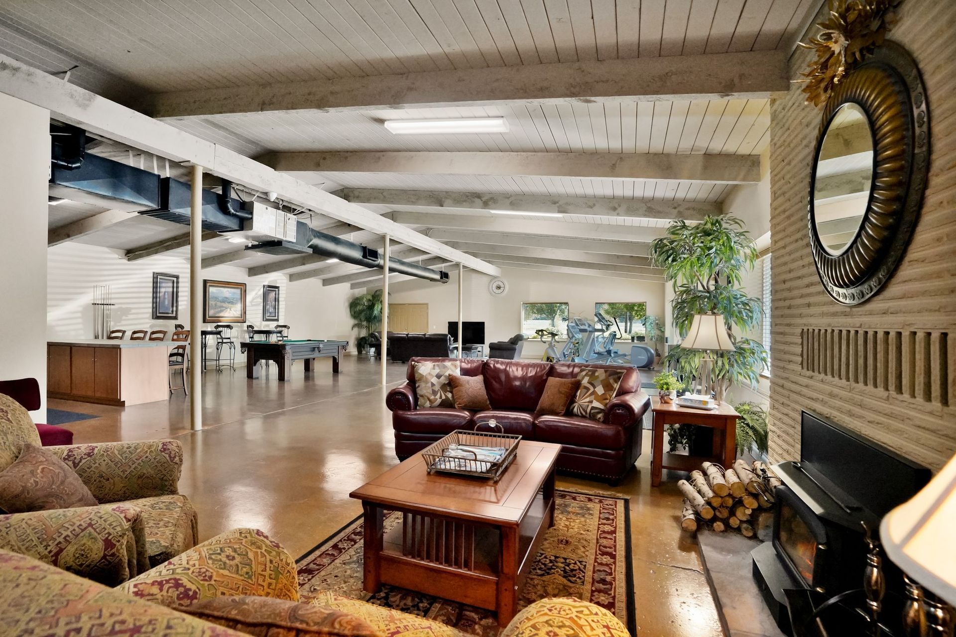 Spacious community lounge with fireplace, sofas, coffee table, bookshelves, exposed beams, and game area in the background.
