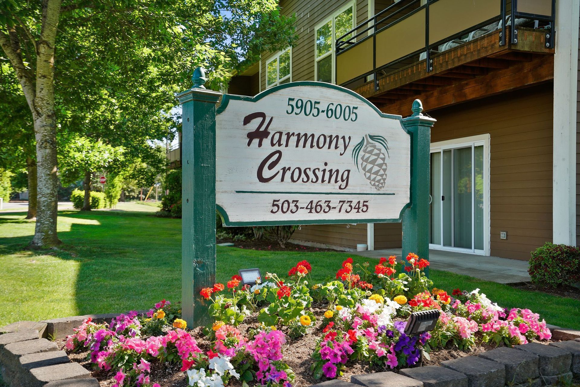 A sign for Harmony Crossing apartments with a phone number, set in a sunny garden with colorful flowers.