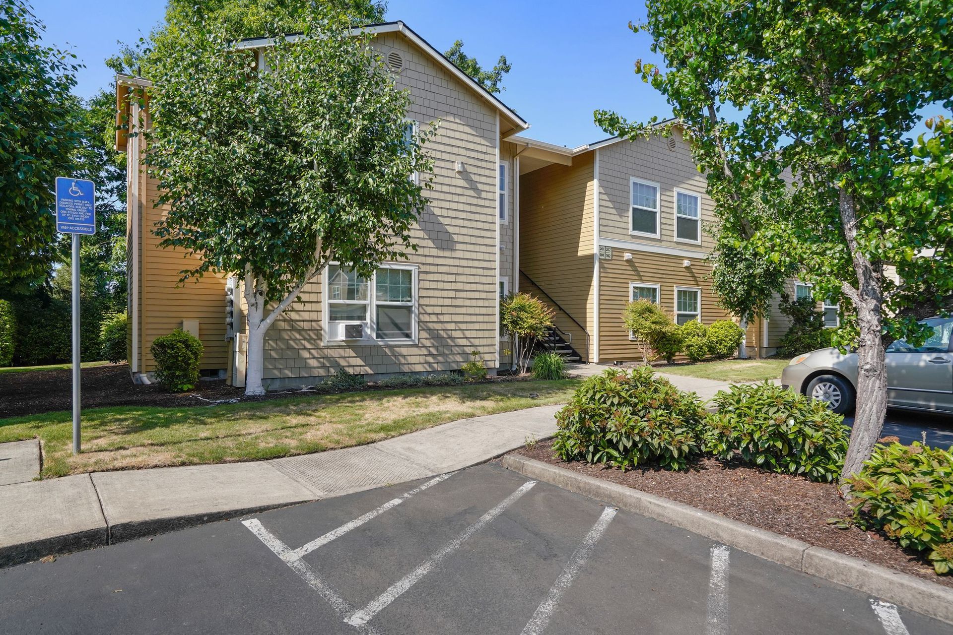 A tan two-story apartment building with a sidewalk, parking spaces, and surrounding trees under a clear blue sky.