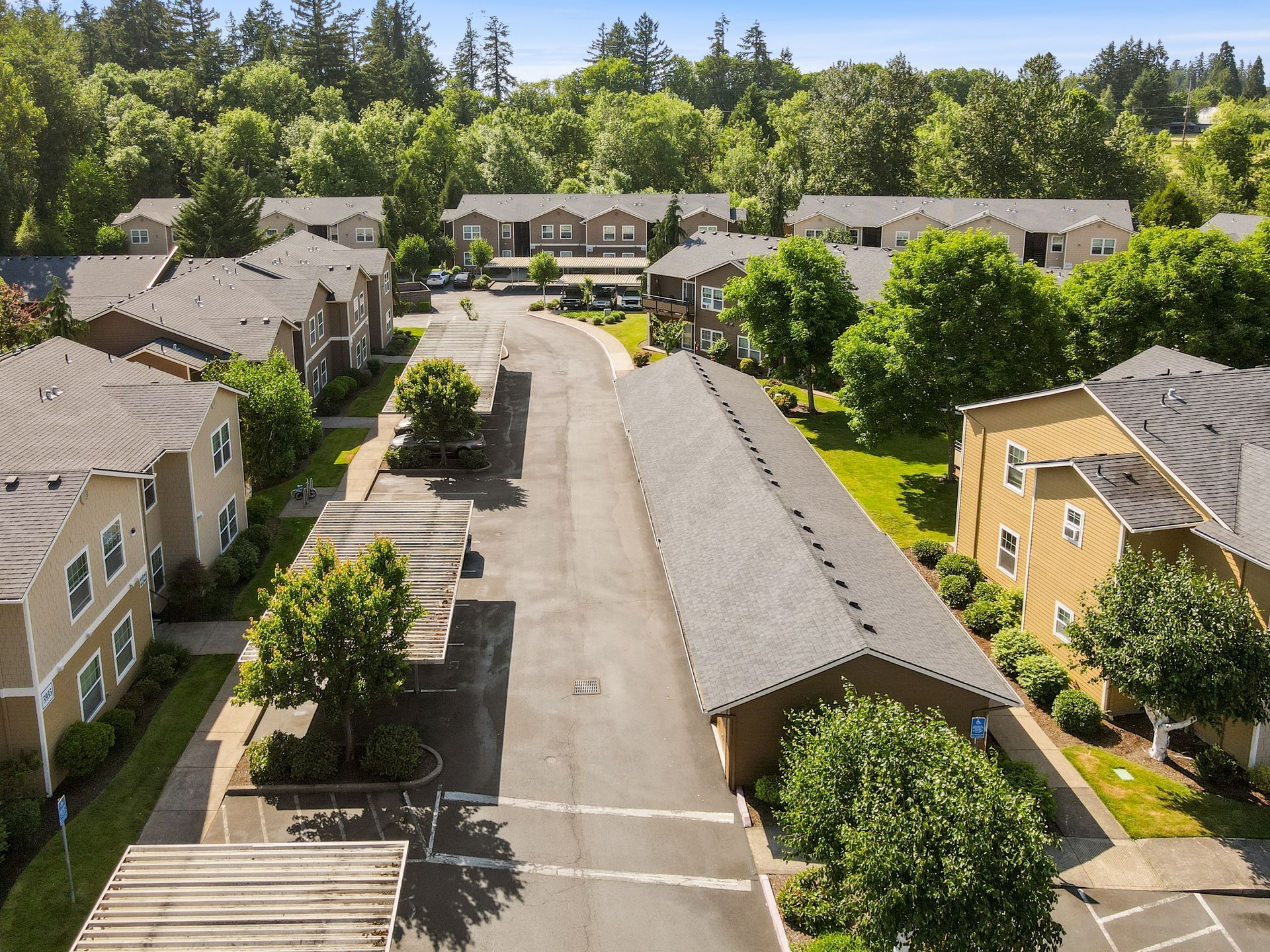 Aerial view of an apartment complex with tan buildings, gray shingled roofs, covered parking, and surrounding trees.
