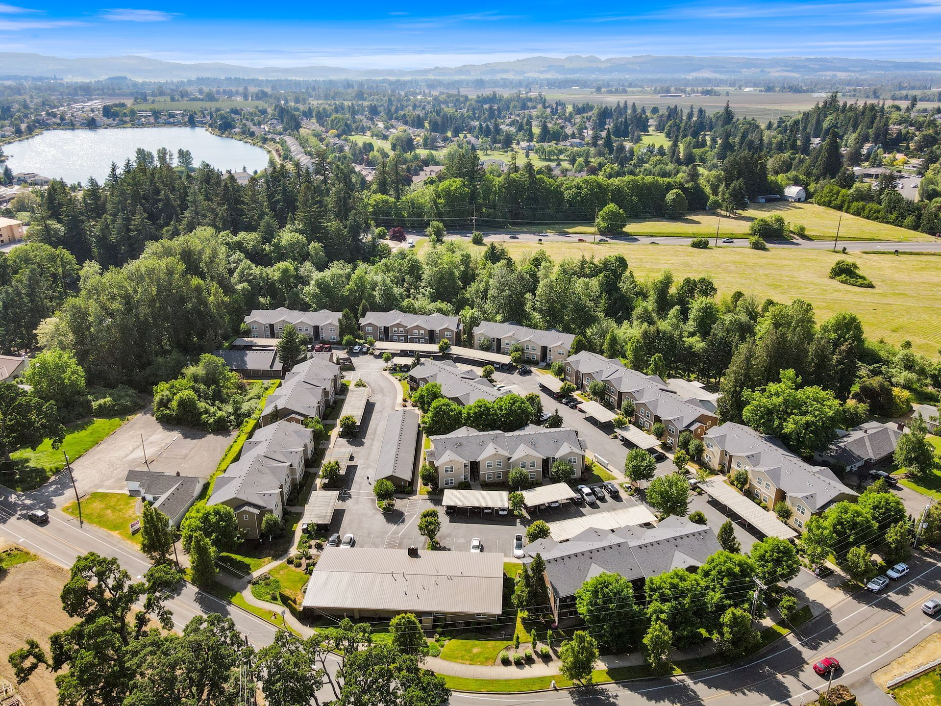 An aerial view of a multi-building apartment complex surrounded by trees, a lake, and grassy fields under a blue sky.