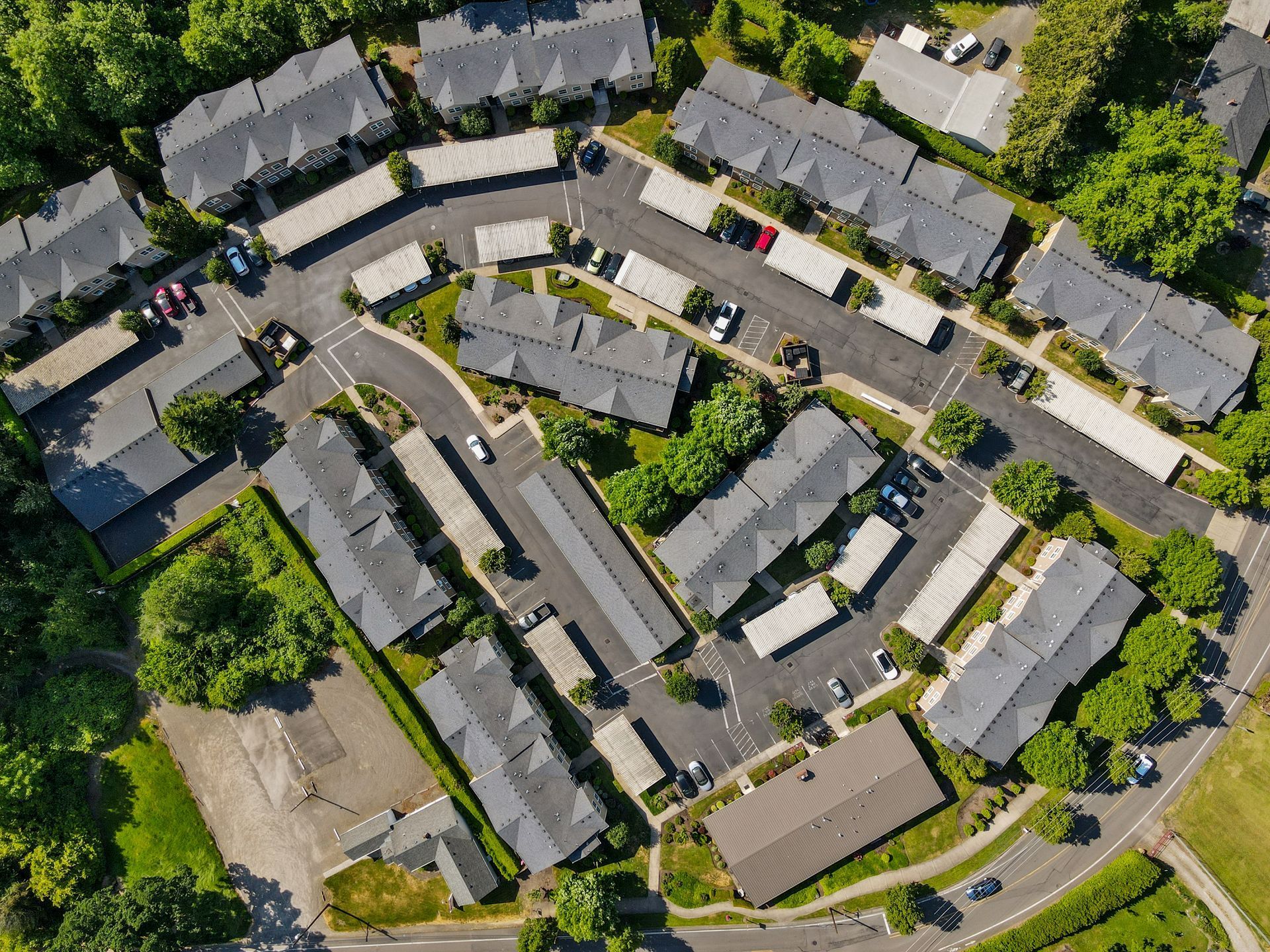 An aerial view of a multi-unit residential apartment complex with grey rooftops, paved parking, and surrounding trees.