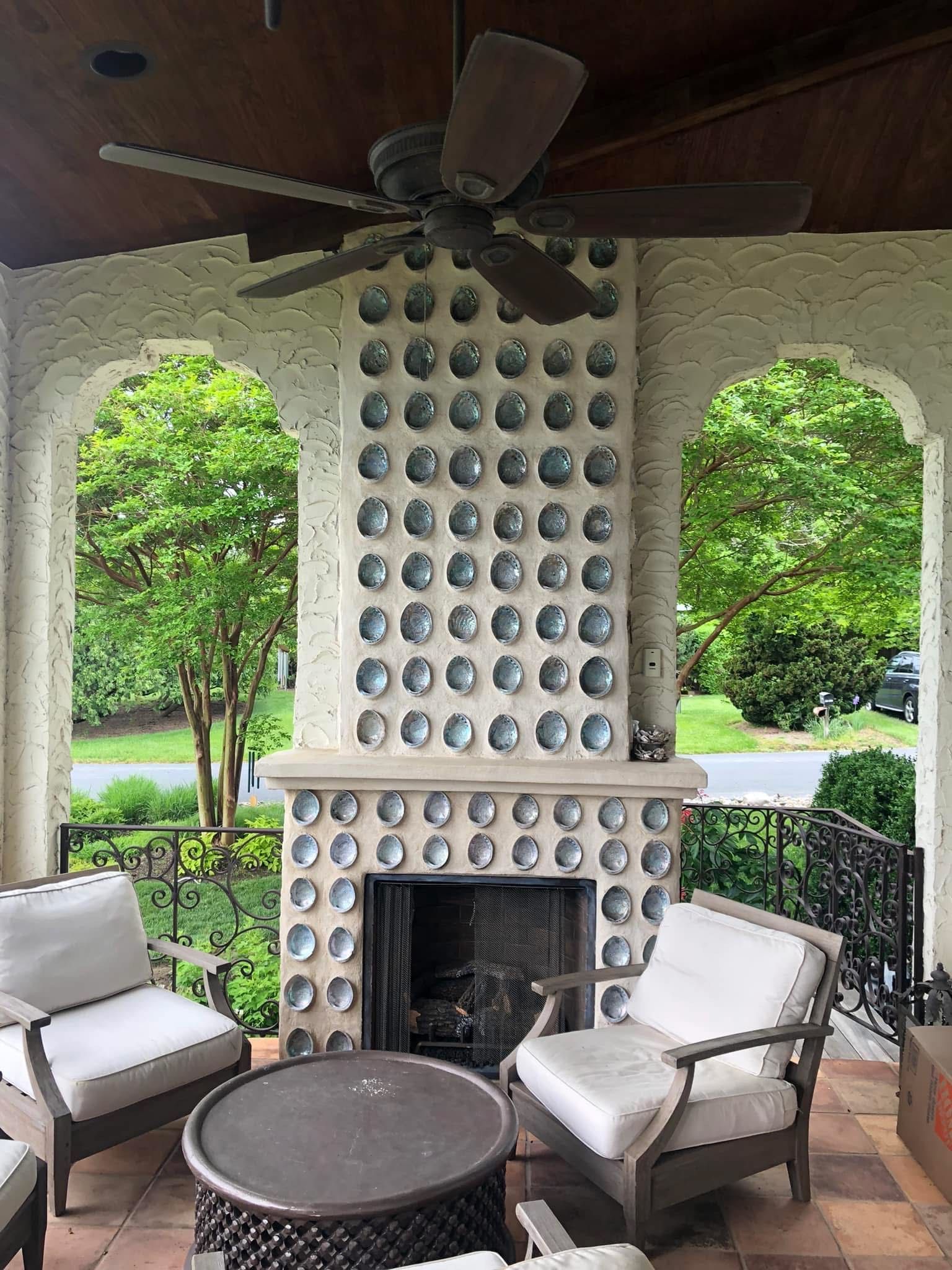 Outdoor patio with fireplace, two armchairs, and a round table. Green trees visible through arched openings.