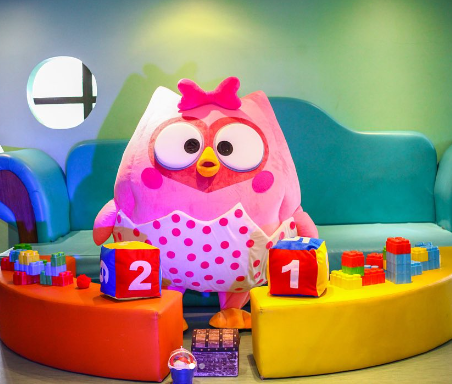 Um mascote coruja rosa está sentado em um sofá cercado por brinquedos