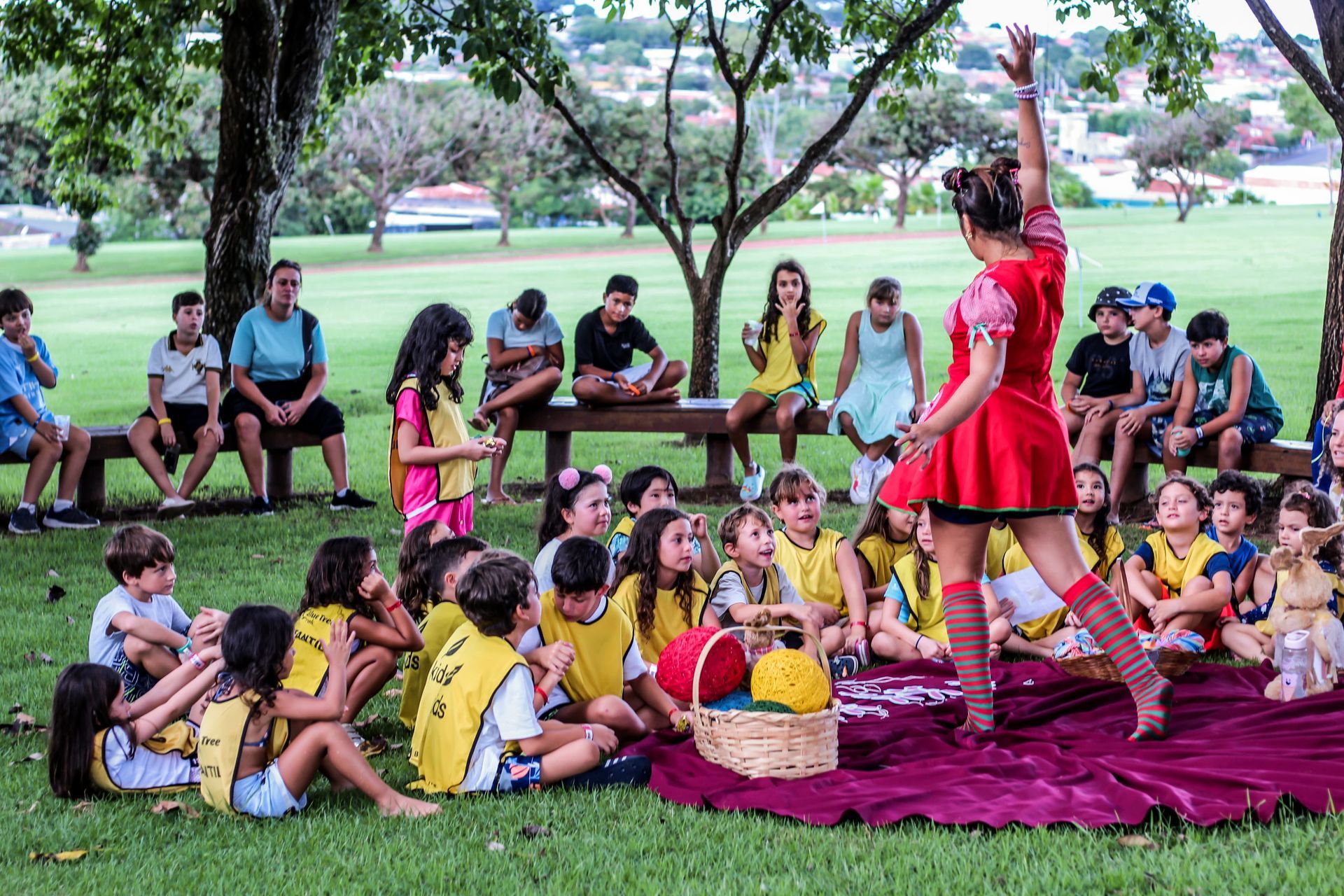 Um grupo de crianças está sentado na grama em um parque assistindo a uma apresentação.