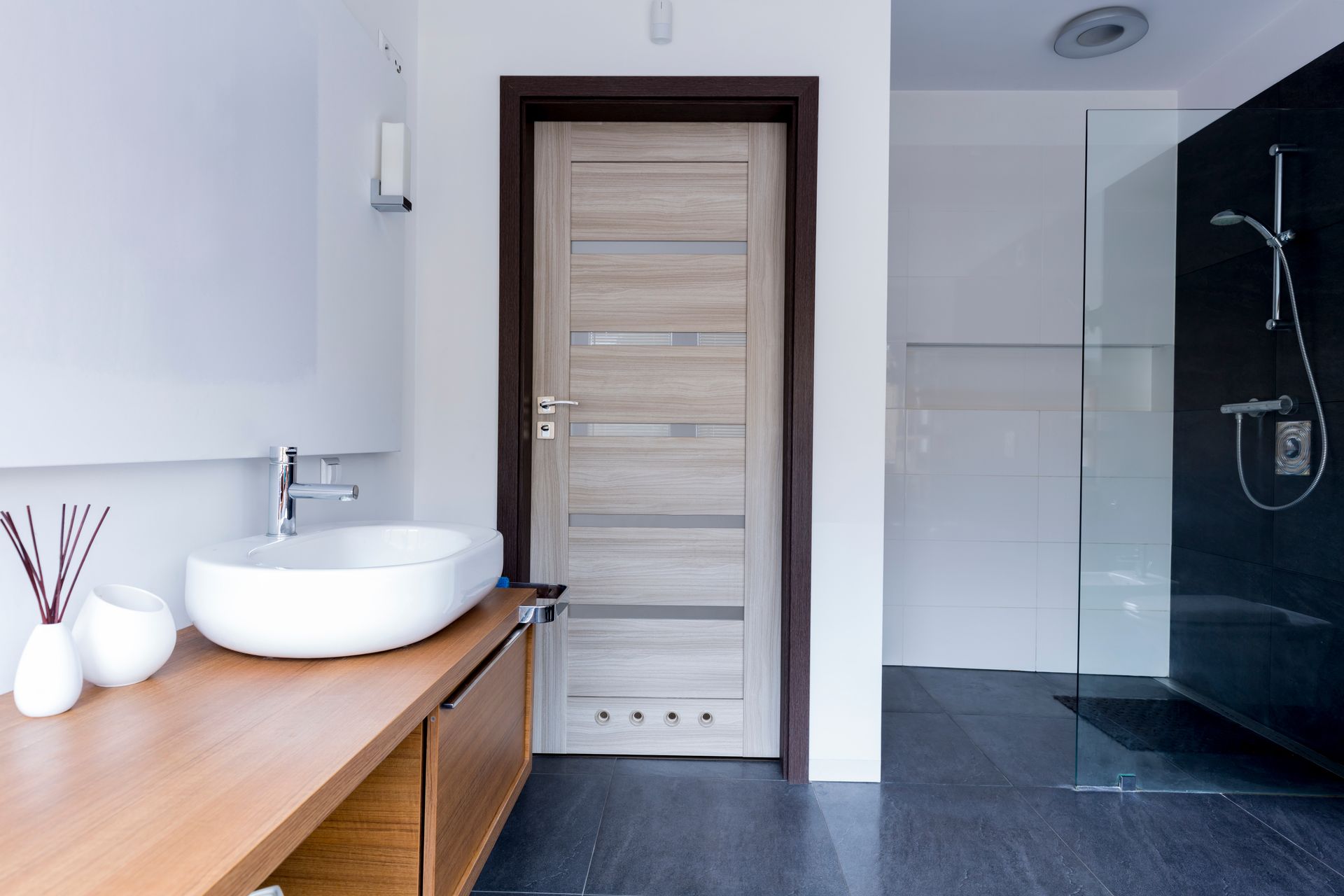 Modern bathroom with wooden vanity, white sink, beige door, and dark shower area Modern bathroom with wooden vanity, white sink, beige door, and dark shower area