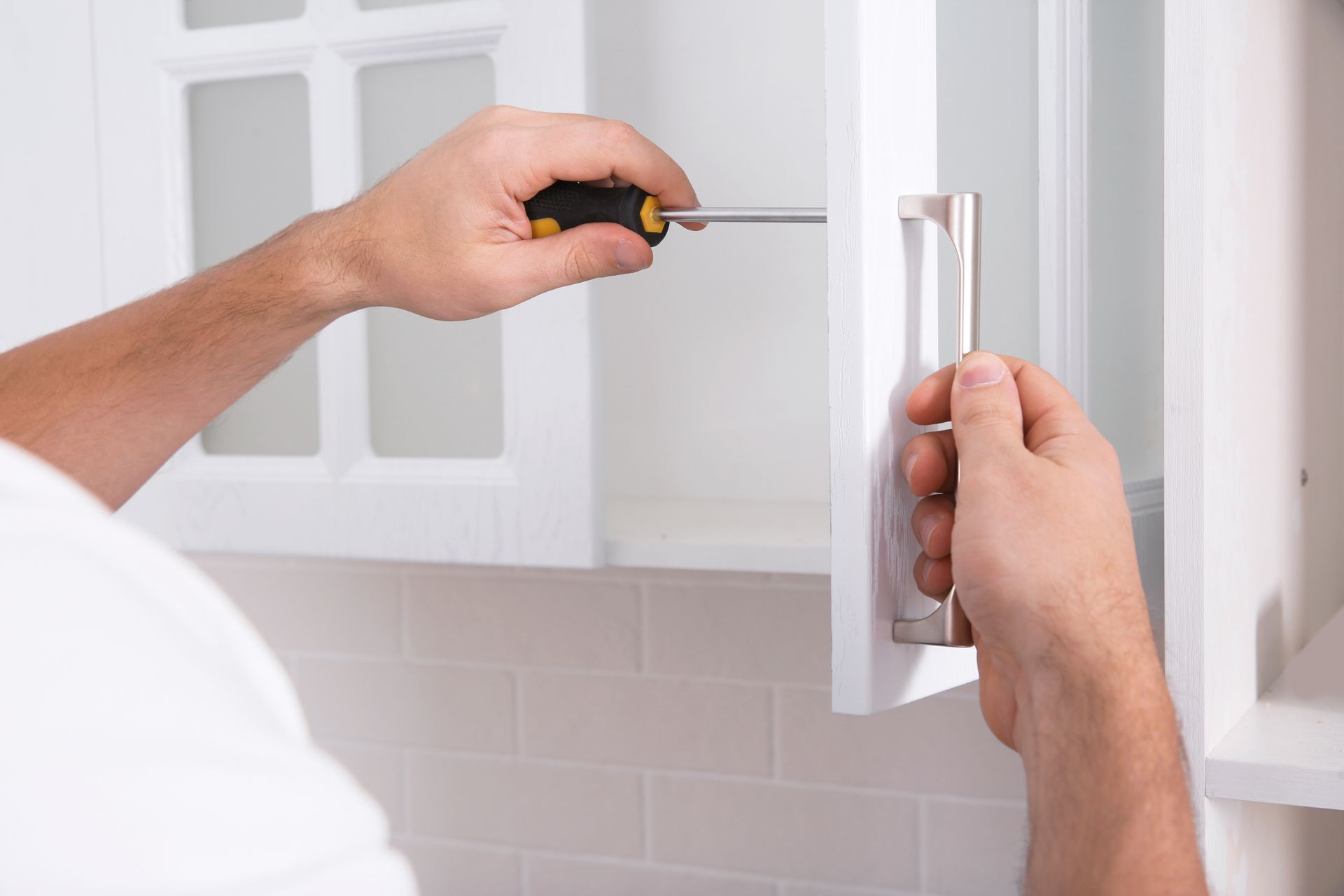 Hands using a screwdriver to install a white cabinet handle in a kitchen. Hands using a screwdriver to install a white cabinet handle in a kitchen.