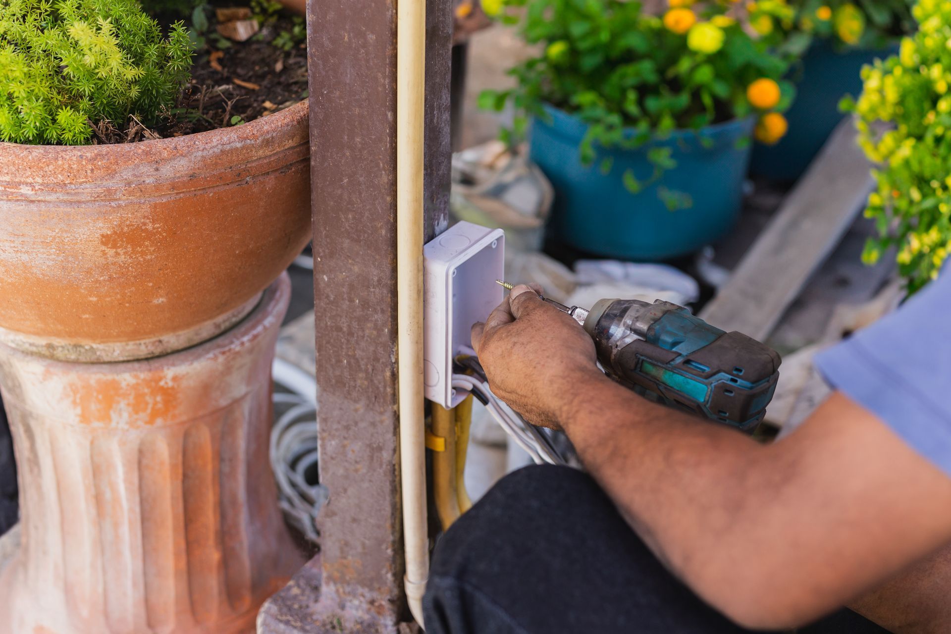 Technician using a power tool to install an outdoor wall device beside potted plants Technician using a power tool to install an outdoor wall device beside potted plants